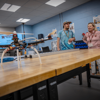 A drone with four propellers sits on a wooden lab table in a classroom. Two instructors stand nearby, talking and smiling, with posters and equipment visible on the walls behind them. A screen in the background displays information about an Explore Makers session.