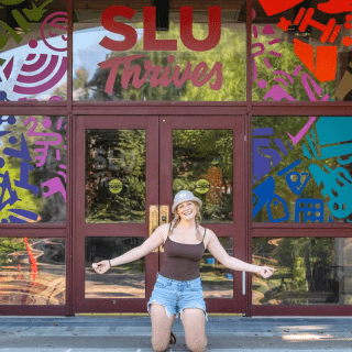 A smiling student kneels with arms open in front of glass doors decorated with colorful graphics and the words “SLU Thrives.” The entrance is framed by brick columns and red Adirondack chairs on both sides. Trees and campus greenery reflect in the glass at Saint Lawrence University.