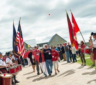 Several Saint Lawrence alums, wearing scarlet and brown apparel, walk through a crowd of cheering people holding flags and people beating drums, during Reunion Weekend.
