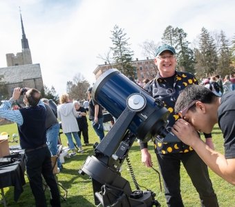 A student peers into a giant black telescope while astronomer Jeff Miller, wearing a black and gold celestial shirt, grins. Gunnison Memorial chapel is in the background. 