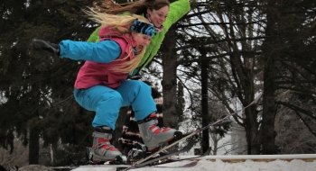 Two students wearing neon snow pants and jackets ski off of a jump together.