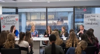 Five panelists in conversation sit at a long table in front of a crowd of students.