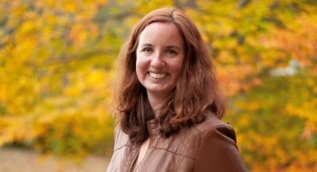 Alanna Gillis, wearing a brown leather jacket, smiles at the camera. She's surrounded by vibrant fall foliage.