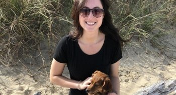 Brittany Hollis sits on a large piece of driftwood holding her small dog at the beach.