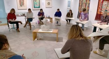 At least 10 students sit in a large circle with large pads of paper in an art gallery as they draw a statue in the middle.