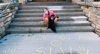 Chloe Mitchell smiles next to sidewalk-chalk writing that says &quot;smile, you are at SLU.&quot;