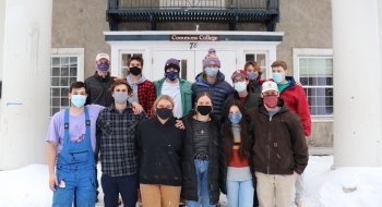 A group of twelve students stand in two rows on the snowy front steps of their residence.