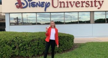 A student in professional attire stands in front of a corporate building with the &quot;Disney University&quot; logo on it.