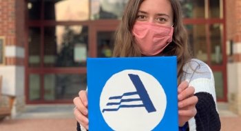 Ella is standing in front of the Saint Lawrence University Student Center and holding a painted canvas with the AmeriCorps logo.