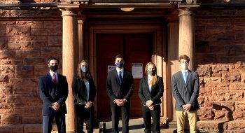 Five students in business clothing stand in front of a sandstone building.