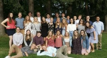 A large group of smiling Saint Lawrence University students gather for a photo in front of a forest on a warm day in early fall.