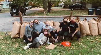Seven girls posing with bags of raked leaves on a grassy lawn.