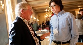 A young man in professional attire talks to his alumni mentor, an older man also in professional attire.