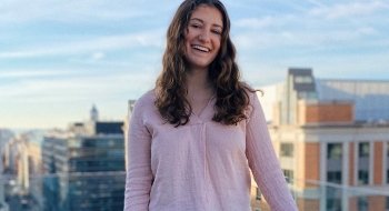 Lily MacCachran wearing a light pink blouse and standing on a balcony overlooking tall buildings in the background.