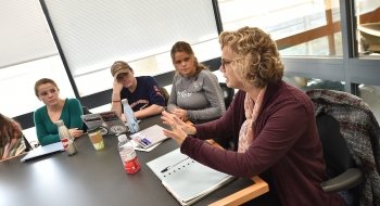A professor sits at the head of a table leading a discussion for students seated around the table.