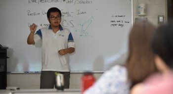 A student stands at a whiteboard at the front of the class explaining a math formula to their audience.