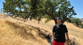 Michael Petroni on a trail through tall yellow grasses with a bristly tree branching out behind him.