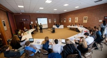 Delegates to the Model United Nations conference are seated at folding tables arranged in a circle around a large screen.