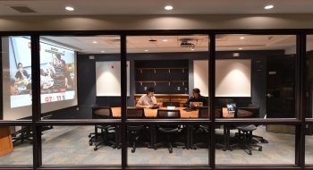 Two students sit at a large conference table in a windowed vestibule in the library.