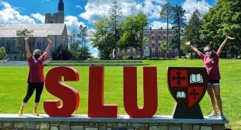 Two students stand next to large foam letters that spell out SLU on a sunny summer day.