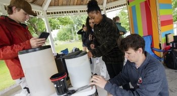 A group of students participating in a photography workshop set up in a gazebo in a park. They’re experimenting with pinhole cameras, which look like large boxes.