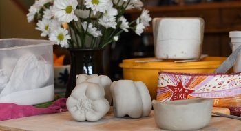 Clay peppers and a clay bowl sit atop a wood cutting board with other tools and kitchen prep items behind them.