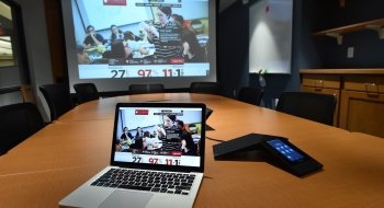 An open laptop sits upon a large conference table in a renovated library space. There's a large projector screen in the background.