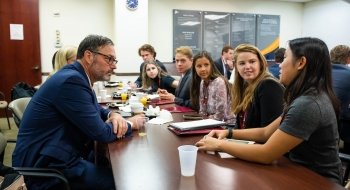 Alumni sit across from current students at a long conference table in a corporate office. A student at the head of a table is asking a question.