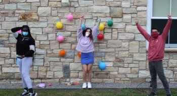 Three students stand in front of a stone wall with their arms in the air. There are colorful balloons behind them.
