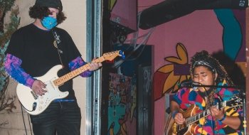 Two students perform at an open mic. One sings and plays guitar and the other plays guitar. Both wear masks.