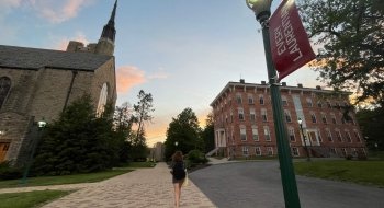 A student walks up a path between a stone church and an academic building on the Saint Lawrence University campus at sunet.