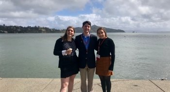 Three students wearing business attire stand in front of a bay on a boardwalk.
