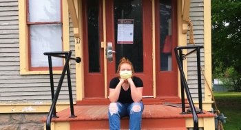 A student, wearing a mask, sits on the front steps of a brightly colored house.