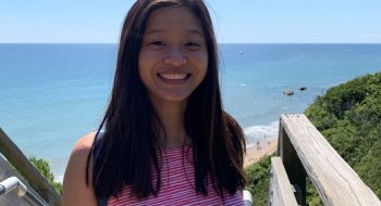 Thea How Choon smiles at the camera. Behind her, there's a beach with a grassy coastline and deep blue ocean water.