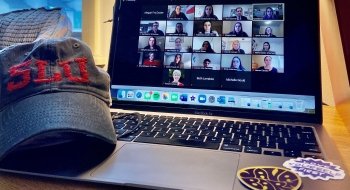 A hat with the letters S, L, U on it next to an open laptop displaying a group of people participating in a networking events.