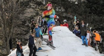 Alex Andrews, wearing a tie dye ski uniform, launches into the air off of a ski jump. A crowd of skiers and snowboarders stand on the jump behind Alex.