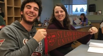 Jesse Meyer and Maddy Schumacher, holding a Saint Lawrence pennant, sit in a classroom. There are other students in the background.