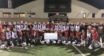 The men's lacrosse team stands together on the field with a check.