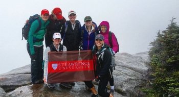 Seven Saint Lawrence students, wearing rain jackets and hats, stand on the summit of a mountain, in the rain, holding a Saint Lawrence University flag.