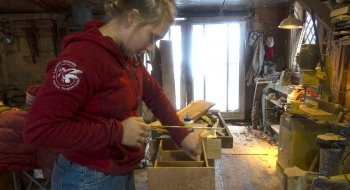 A Saint Lawrence University student, wearing a red hooded sweatshirt, saws into wood in a workshop.
