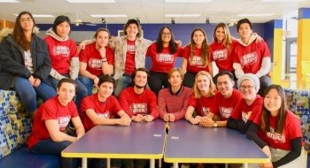 A group of students wearing red shirts sit in two rows among booths in a school cafeteria.