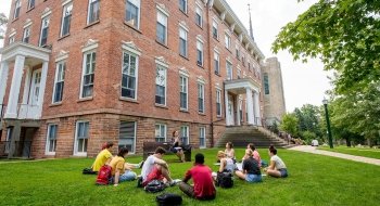Professor Sarah Gates sits on a bench outside of Richardson Hall teaching a class to ten Saint Lawrence University students on a sunny day.