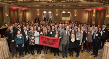 A large group of Saint Lawrence University alumni, wearing professional attire, stand in a group holding a scarlet and brown Saint Lawrence University flag.