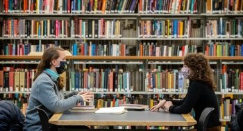 Alanna Gillis, wearing a light blue sweater and a mask, sits at a table in the library across from Alleigh Szabo, who is wearing a black sweater and a mask. In the background, there is a large book shelf filled with colorful books.