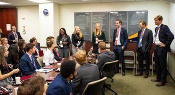 A group of young Saint Lawrence alumni dressed professionally stand at the front of a room and talk with a group of students seated at a long table.