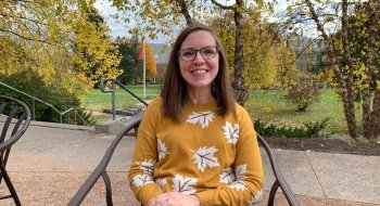 Laura Lavoie, wearing a mustard yellow sweater with a leaf patter, sits in a chair on an outdoor walkway. There are trees with yellow leaves in the background.
