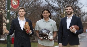 Andrew Matthews, Tyler Grochet, and Ben Barba, wearing business attire and hold athletic equipment, stand next to one another on a campus walkway on an overcast day.