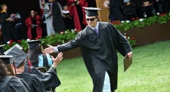 A Saint Lawrence student, wearing graduation attire and sunglasses. high-fives a fellow member of the graduating class.