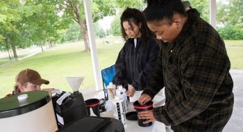 Sarath Novas sets up photography equipment in a gazebo in a park. They’re experimenting with pinhole cameras, which look like large boxes.