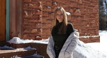 A student wearing a black sweater and a white puffy coat stands in front of a sandstone brick building on a sunny, snow-covered winter day.